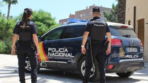 two spanish police officers, one male and one female, stand in front of their police car