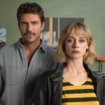A manin a grey shirt and woman in a yellow and blue striped top pose for a promotional photo in front of a green wall.